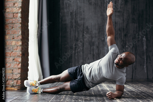 Strong and muscular smiling African American doing a side bar while standing on his left arm and leg with his right arm raised.