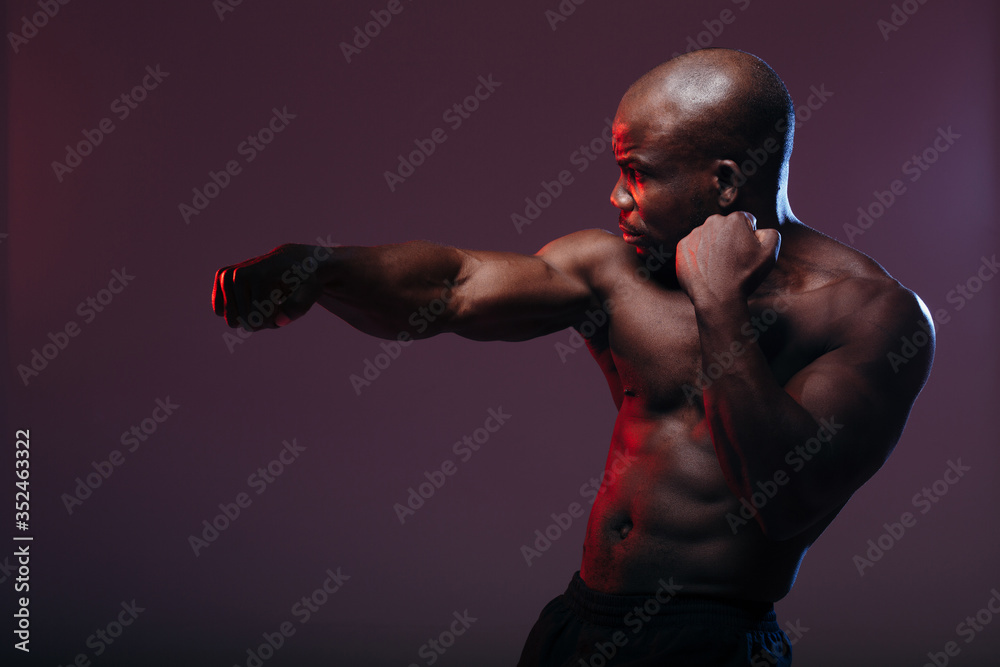 A dark-skinned boxer is training with a naked torso. The athlete with embossed muscles is backlit with red light.