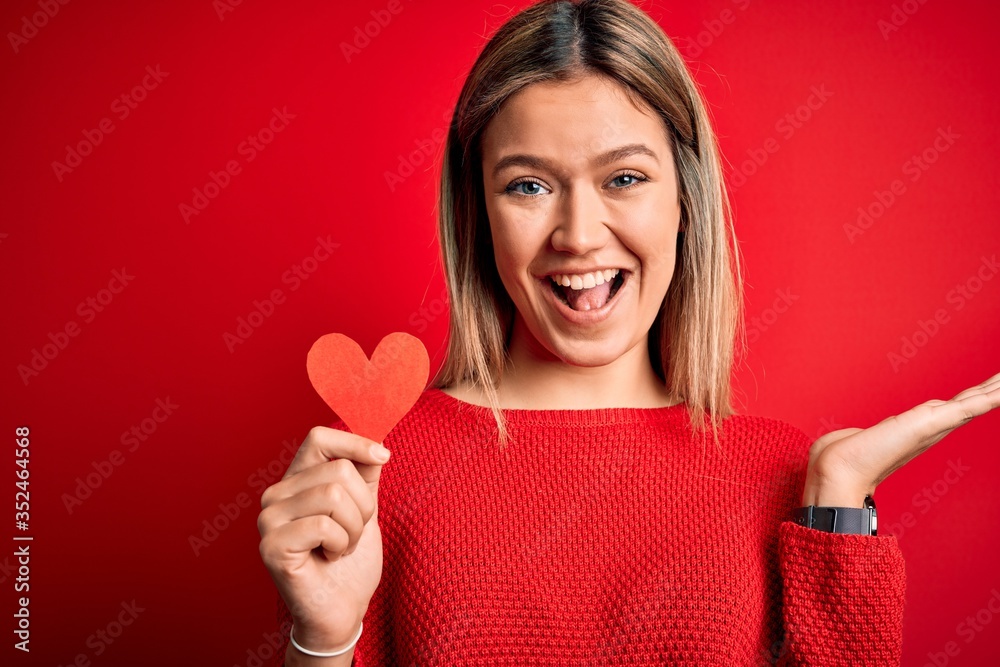 Young beautiful woman holding heart card standing over isolated red background very happy and excited, winner expression celebrating victory screaming with big smile and raised hands Young beautiful woman holding heart card standing over isolated red background very happy and excited, winner expression celebrating victory screaming with big smile and raised hands