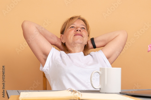 Middle age woman in white t-shirt sitting at the table, holds her hands behinde her head and shows hairy unshaven female armpits photo