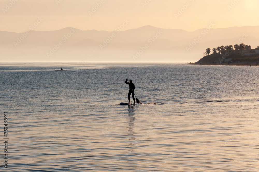 A man with an oar stands on a board in the sea on a background of mountains and palm trees during sunset
