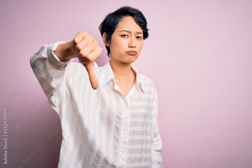 Young beautiful asian girl wearing casual shirt standing over isolated pink background looking unhappy and angry showing rejection and negative with thumbs down gesture. Bad expression. Young beautiful asian girl wearing casual shirt standing over isolated pink background looking unhappy and angry showing rejection and negative with thumbs down gesture. Bad expression.