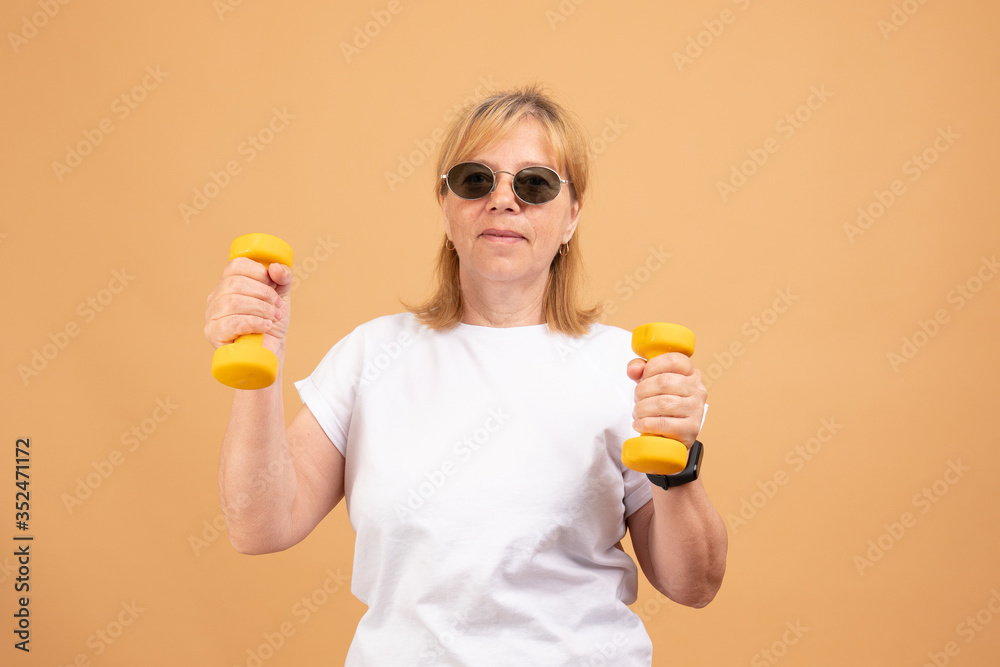 Active energetic happy elderly Caucasian female with blonde hair enjoying physical exercises indoors, training at home using dumbbells