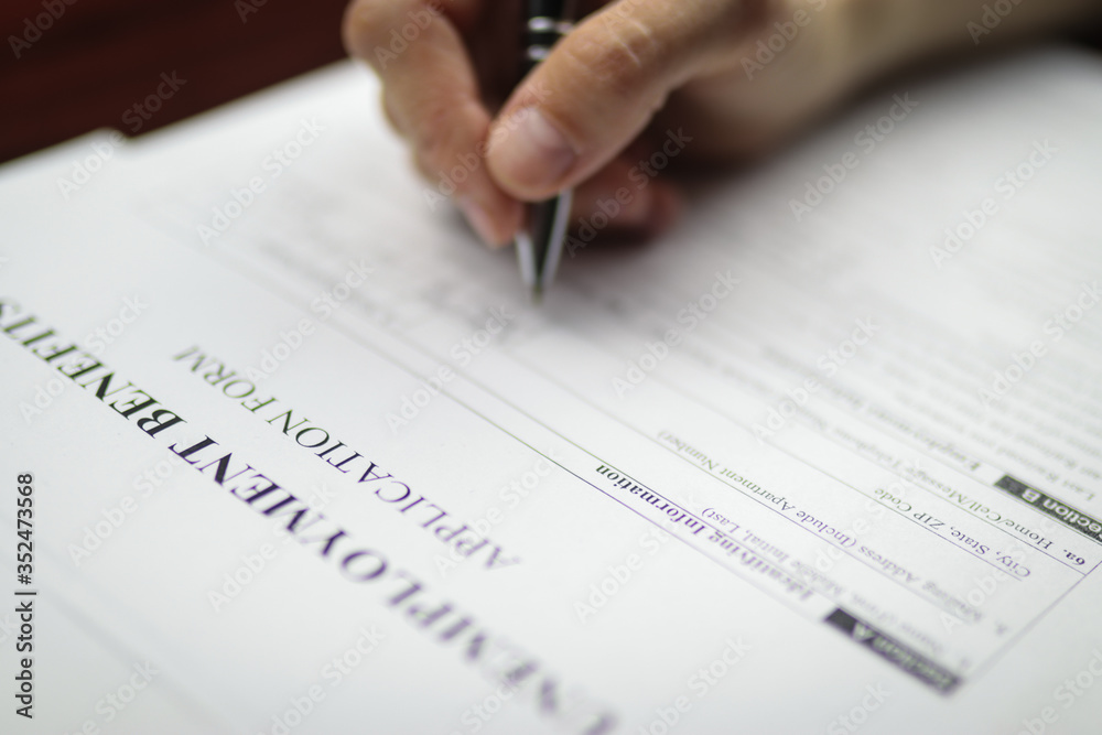 Close-up of a person's hand filing social security benefits application form. Concept of Covid-19 coronavirus and stay at home order impact on economy, world economic crisis, unemployment. Close-up of a person's hand filing social security benefits application form. Concept of Covid-19 coronavirus and stay at home order impact on economy, world economic crisis, unemployment.