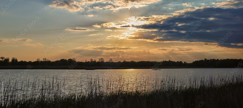 Panoramic photo of beautiful sunset that hide behind clouds with blue sky trees and water in the foreground