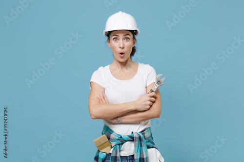 Amazed young woman in protective helmet hardhat hold adjustable wrench isolated on blue background. Instruments accessories for renovation apartment room. Repair home concept. Holding hands crossed.