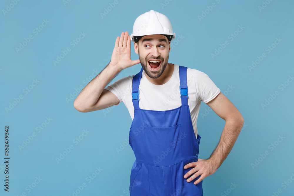 Excited young man in coveralls protective helmet hardhat isolated on blue background. Instruments accessories for renovation apartment room. Repair home concept. Try to hear you with hand near ear. Excited young man in coveralls protective helmet hardhat isolated on blue background. Instruments accessories for renovation apartment room. Repair home concept. Try to hear you with hand near ear.