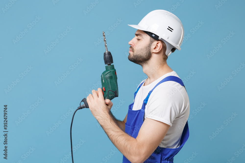 Side view of young man in coveralls protective helmet hardhat hold electric drill isolated on pastel blue background studio. Instruments accessories for renovation apartment room. Repair home concept. Side view of young man in coveralls protective helmet hardhat hold electric drill isolated on pastel blue background studio. Instruments accessories for renovation apartment room. Repair home concept.