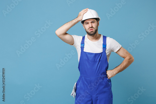 Exhausted preoccupied young man in coveralls protective helmet hardhat isolated on blue background studio. Instruments accessories for renovation apartment room. Repair home concept. Put hand on head.