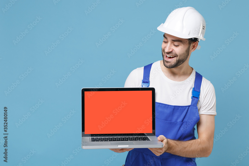 Funny man in coveralls protective helmet hardhat hold laptop computer with blank empty screen isolated on blue background. Instruments accessories for renovation apartment room. Repair home concept. Funny man in coveralls protective helmet hardhat hold laptop computer with blank empty screen isolated on blue background. Instruments accessories for renovation apartment room. Repair home concept.