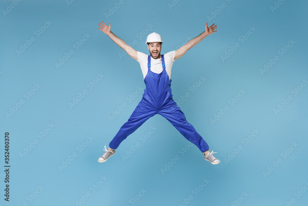 Excited young man in coveralls protective helmet hardhat isolated on blue background. Instruments accessories for renovation apartment room. Repair home concept. Jumping, spreading hands and legs.