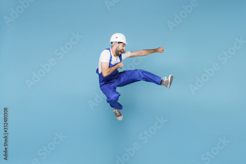 Funny young man in coveralls protective helmet hardhat isolated on blue background in studio. Instruments accessories for renovation apartment room. Repair home concept. Having fun, jumping fighting.