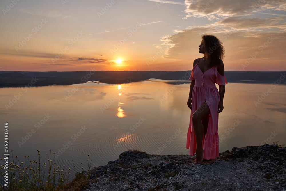 Girl in long pink dress on rock over lake in evening.