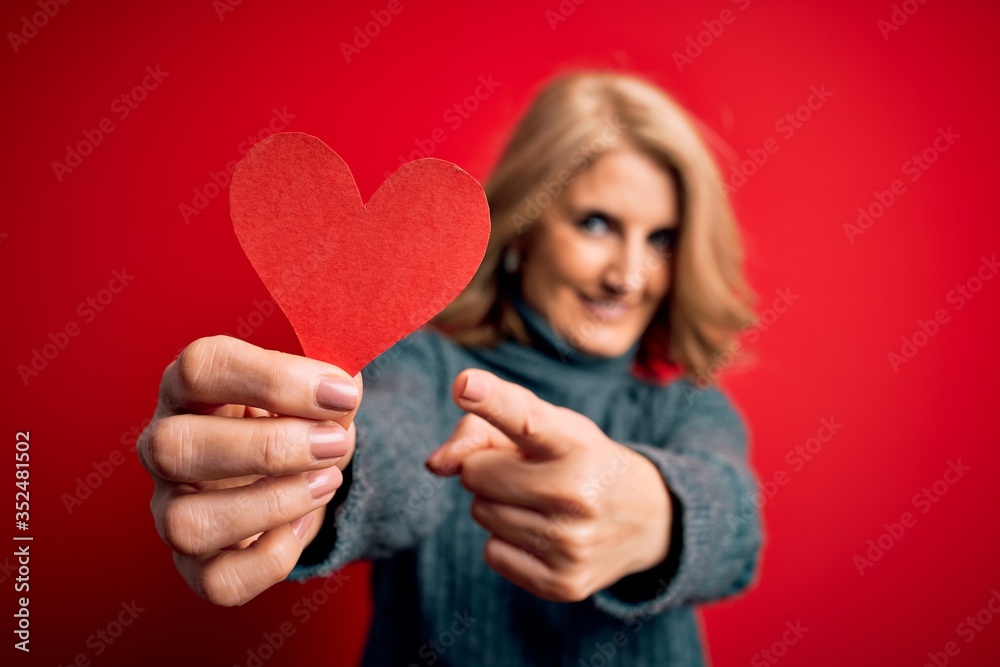 Middle age beautiful blonde romantic woman holding red paper heart celebrating valentine day very happy pointing with hand and finger Middle age beautiful blonde romantic woman holding red paper heart celebrating valentine day very happy pointing with hand and finger