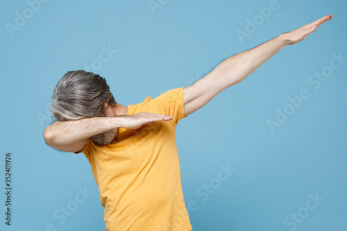 Elderly gray-haired mustache bearded man in casual yellow t-shirt posing isolated on pastel blue background studio portrait. People lifestyle concept. Mock up copy space. Showing DAB dance gesture.