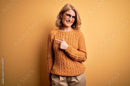 Middle age beautiful blonde woman wearing casual sweater and glasses over yellow background cheerful with a smile on face pointing with hand and finger up to the side with happy and natural expression