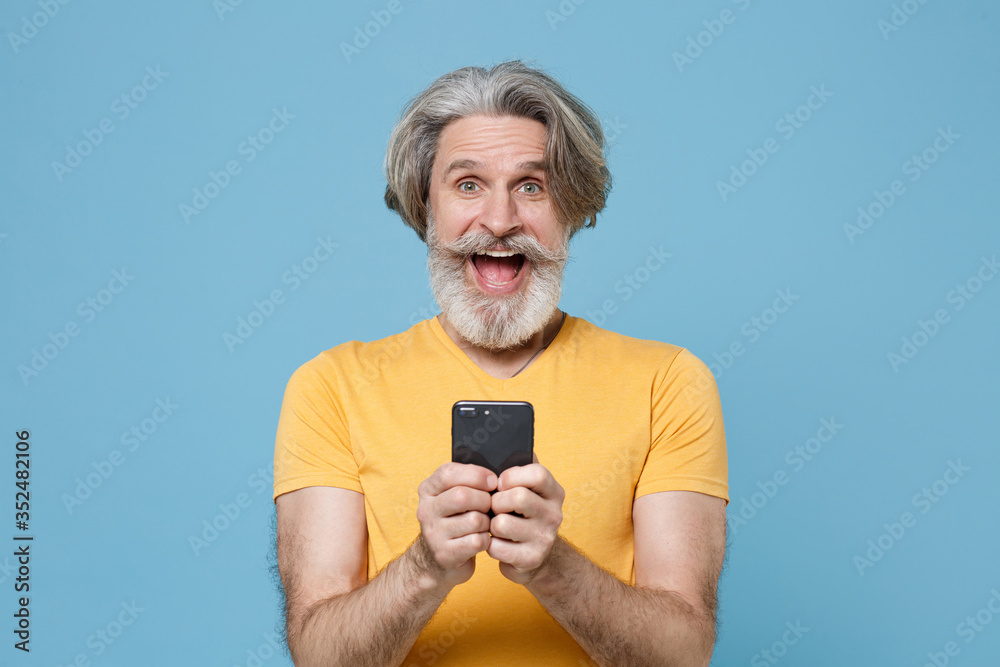 Surprised elderly gray-haired mustache bearded man in casual yellow t-shirt posing isolated on blue background. People lifestyle concept. Mock up copy space. Using mobile phone, typing sms message. Surprised elderly gray-haired mustache bearded man in casual yellow t-shirt posing isolated on blue background. People lifestyle concept. Mock up copy space. Using mobile phone, typing sms message.