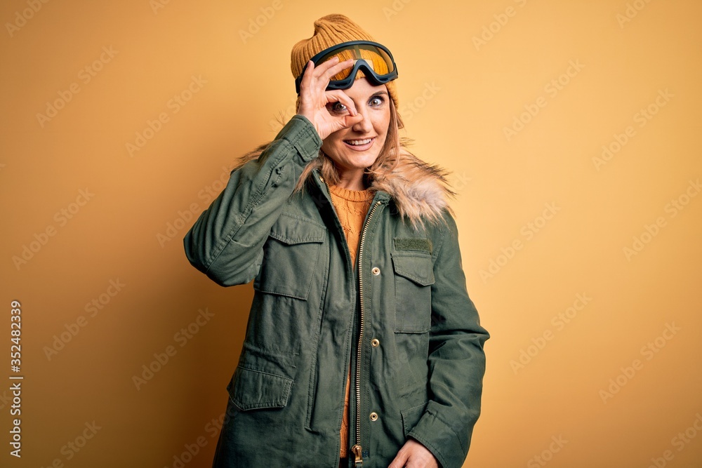 Middle age beautiful blonde skier woman wearing snow sportwear and ski goggles doing ok gesture with hand smiling, eye looking through fingers with happy face.