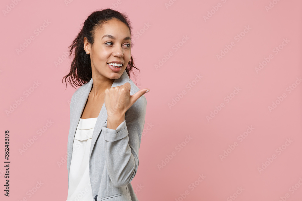 Side view of smiling young african american business woman in grey suit white shirt posing isolated on pink background studio portrait. Achievement career wealth business concept. Point thumb aside.