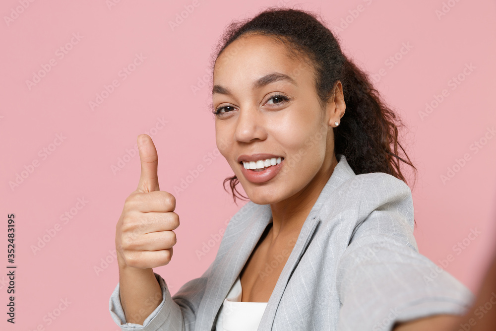 Close up of young african american business woman in grey suit white shirt isolated on pink background. Achievement career wealth business concept. Doing selfie shot on mobile phone, showing thumb up. Close up of young african american business woman in grey suit white shirt isolated on pink background. Achievement career wealth business concept. Doing selfie shot on mobile phone, showing thumb up.
