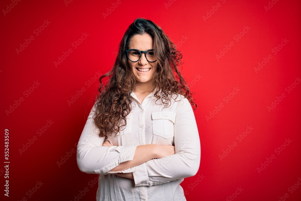 Young beautiful woman with curly hair wearing shirt and glasses over red background happy face smiling with crossed arms looking at the camera. Positive person. Young beautiful woman with curly hair wearing shirt and glasses over red background happy face smiling with crossed arms looking at the camera. Positive person.