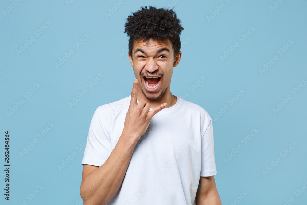Crazy young african american guy in casual white t-shirt posing isolated on pastel blue background. People lifestyle concept. Mock up copy space. Depicting heavy metal rock sign, horns up gesture. Crazy young african american guy in casual white t-shirt posing isolated on pastel blue background. People lifestyle concept. Mock up copy space. Depicting heavy metal rock sign, horns up gesture.