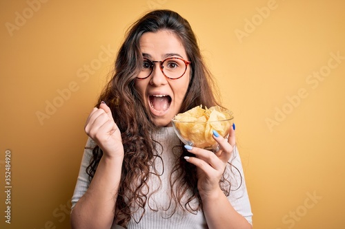 Young beautiful woman with curly hair holding bowl of chips potatoes over yellow background screaming proud and celebrating victory and success very excited  cheering emotion