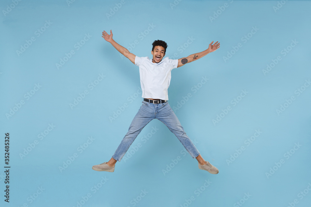 Excited young african american guy in casual white t-shirt posing isolated on pastel blue background studio portrait. People lifestyle concept. Mock up copy space. Jumping, spreading hands and legs.