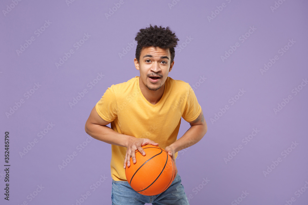 Young african american guy basketball player in casual yellow t-shirt posing isolated on pastel violet background studio. People emotions, sport leisure lifestyle concept. Play basketball with ball.