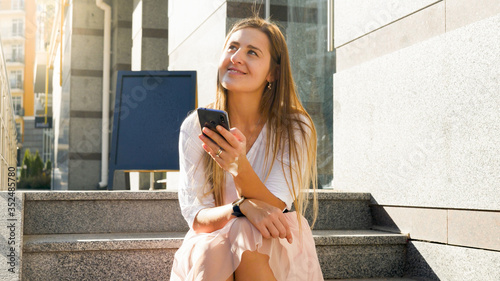 Portrait of smiling dreamy young woman sitting on the stone stairs on city street and holding mobile phone