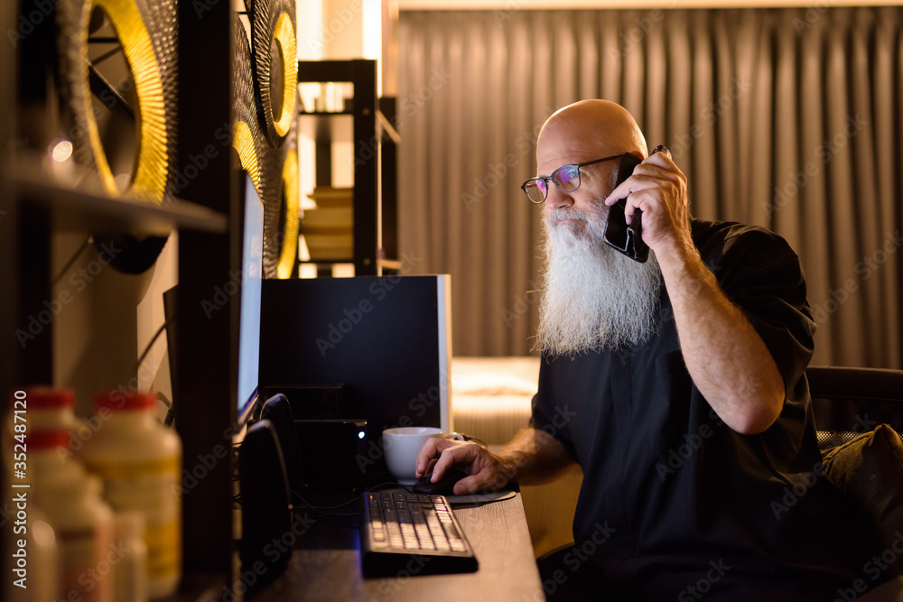 Mature bald bearded man talking on the phone while working overtime at home late at night