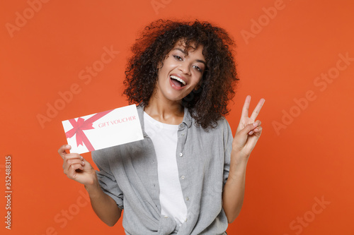 Funny young african american woman girl in gray casual clothes isolated on orange background studio portrait. People lifestyle concept. Mock up copy space. Hold gift certificate, showing victory sign.