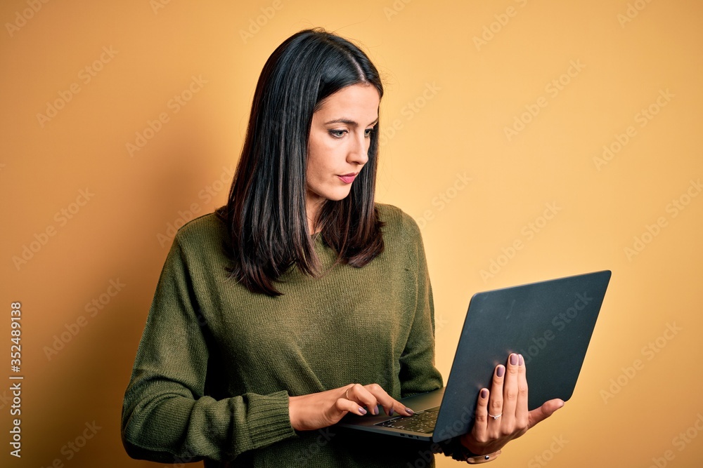 Young brunette woman with blue eyes working using computer laptop over yellow background Checking the time on wrist watch, relaxed and confident