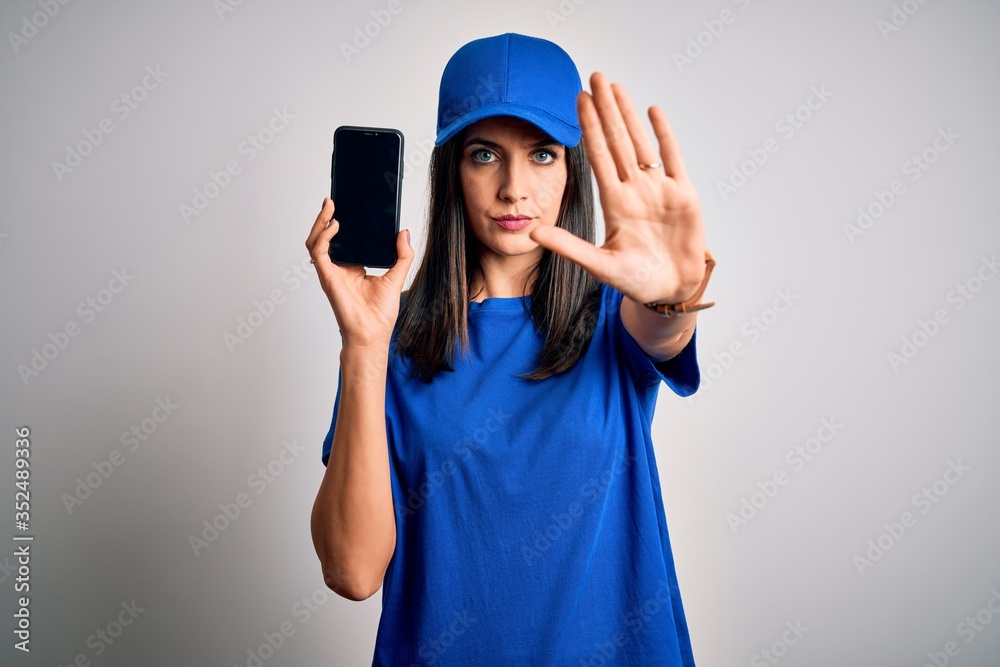 Young delivery woman with blue eyes wearing cap holding smartphone with open hand doing stop sign with serious and confident expression, defense gesture Young delivery woman with blue eyes wearing cap holding smartphone with open hand doing stop sign with serious and confident expression, defense gesture