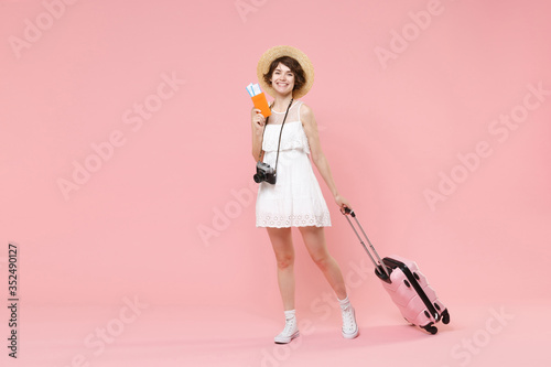 Smiling tourist girl in dress hat with suitcase photo camera isolated on pink background. Traveling abroad to travel weekends getaway. Air flight journey concept. Hold passport tickets boarding pass. © ViDi Studio