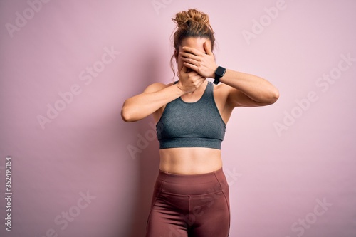 Young beautiful blonde sportswoman doing sport wearing sportswear over pink background Covering eyes and mouth with hands, surprised and shocked. Hiding emotion © Krakenimages.com