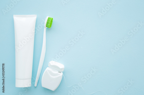 White tube of toothpaste, toothbrush with green bristles and container of dental floss on pastel blue background. People teeth hygiene concept. Empty place for text or logo. Closeup. Top down view.