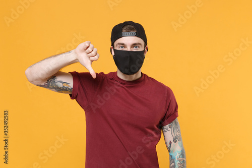 Young tattooed man guy in casual t-shirt cap black face mask posing isolated on yellow background studio portrait. People sincere emotions lifestyle concept. Mock up copy space. Showing thumb down.