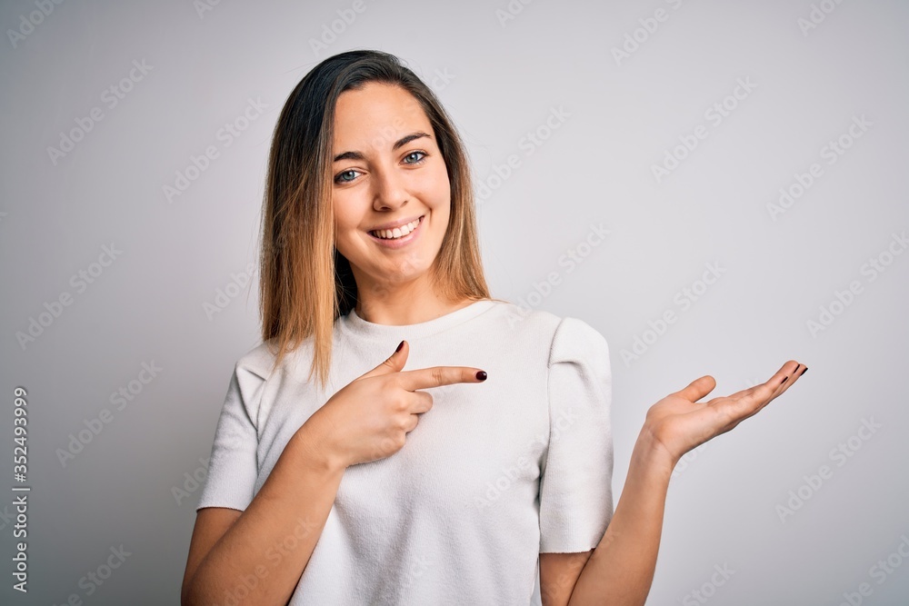 Young beautiful blonde woman with blue eyes wearing casual t-shirt over white background amazed and smiling to the camera while presenting with hand and pointing with finger. Young beautiful blonde woman with blue eyes wearing casual t-shirt over white background amazed and smiling to the camera while presenting with hand and pointing with finger.