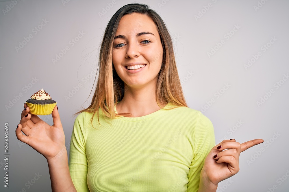 Young beautiful woman with blue eyes eating sweet chocolate cupcake over white background very happy pointing with hand and finger to the side Young beautiful woman with blue eyes eating sweet chocolate cupcake over white background very happy pointing with hand and finger to the side