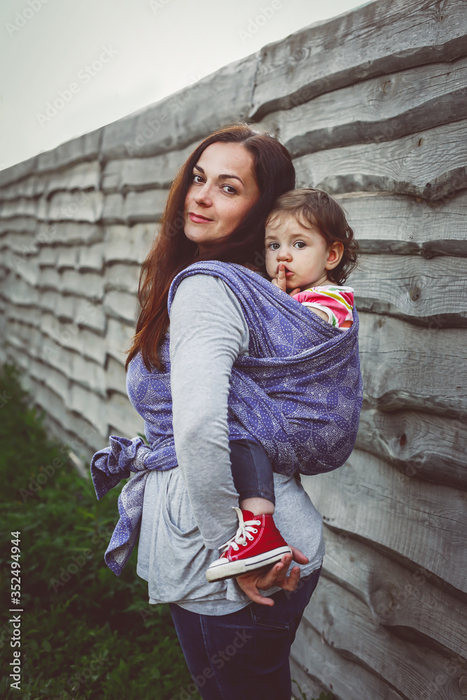 Mother hold baby girl back in sling wrap carrier at rural wooden fence background, cute female child and active modern mom