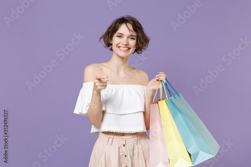 Smiling young woman girl in summer clothes hold package bag with purchases isolated on pastel violet background. Shopping discount sale concept. Mock up copy space. Pointing index finger on camera.
