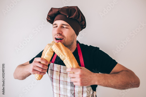 Young man isolated over background. Guy in cook uniform biting two pieces of french baguette and enoy it. Eating fresh tasty delicious french bread. photo