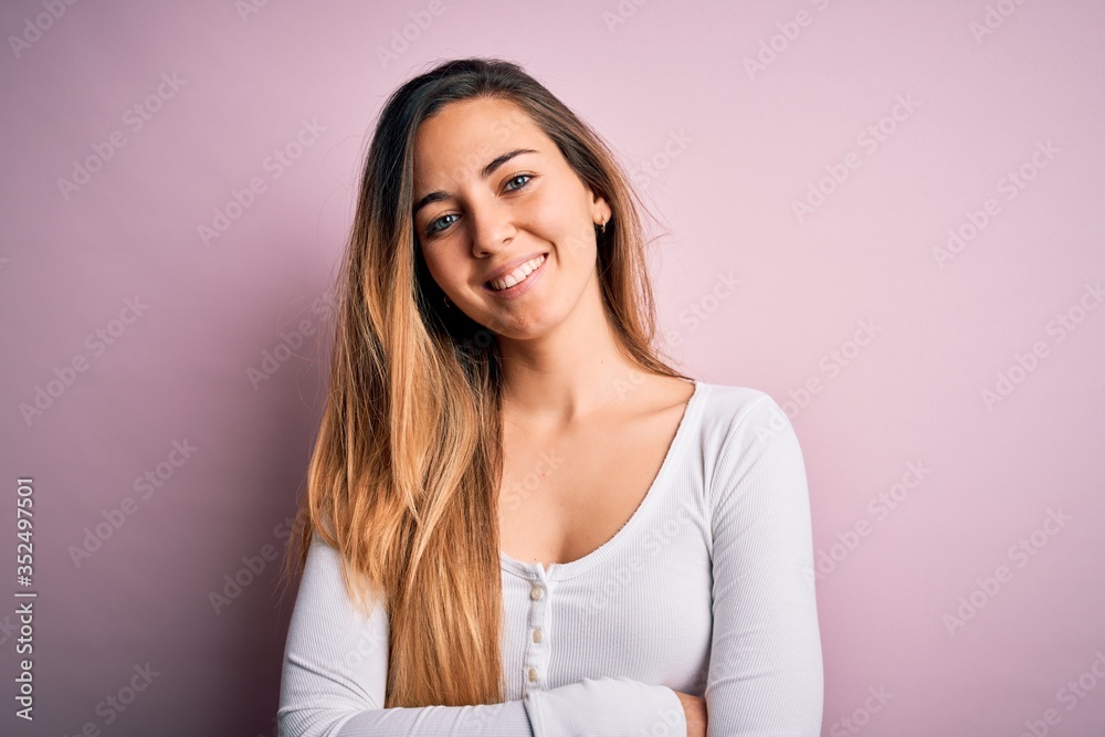Young beautiful blonde woman with blue eyes wearing white t-shirt over pink background happy face smiling with crossed arms looking at the camera. Positive person. Young beautiful blonde woman with blue eyes wearing white t-shirt over pink background happy face smiling with crossed arms looking at the camera. Positive person.