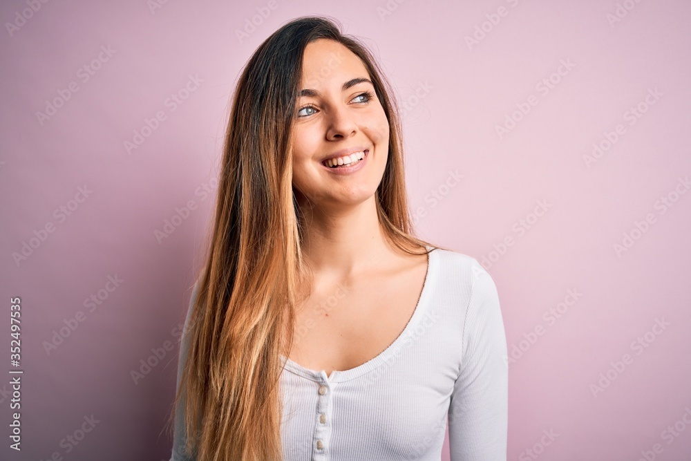 Fototapeta premium Young beautiful blonde woman with blue eyes wearing white t-shirt over pink background looking away to side with smile on face, natural expression. Laughing confident.