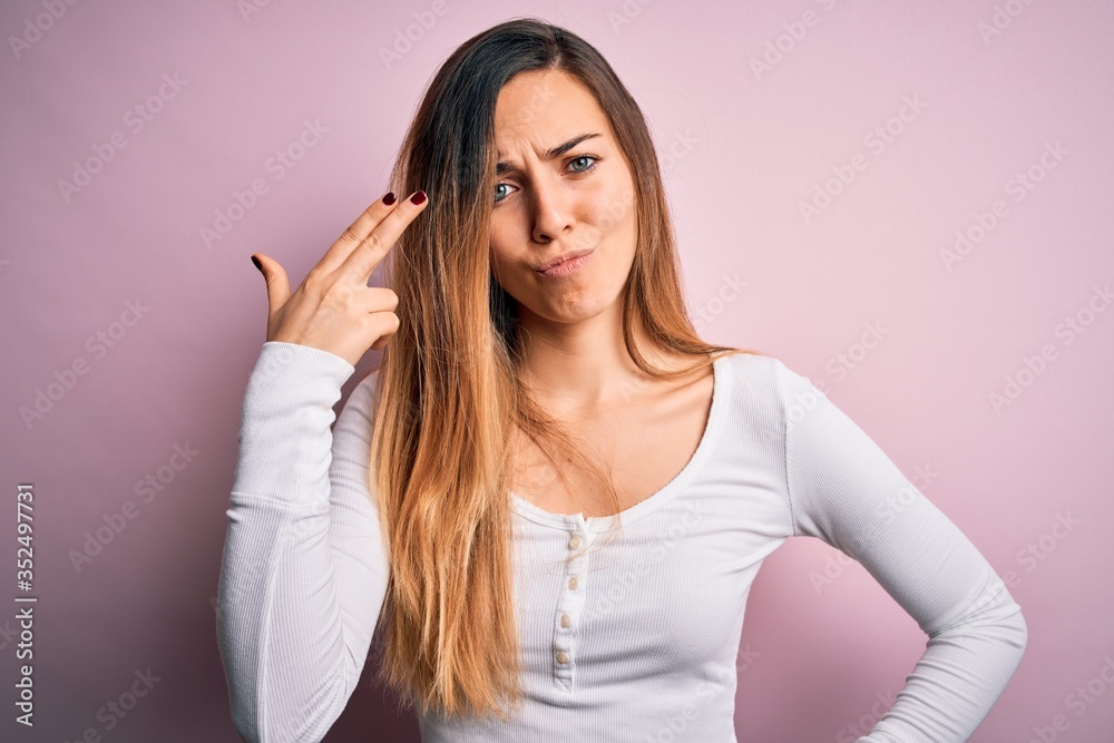 Young beautiful blonde woman with blue eyes wearing white t-shirt over pink background Shooting and killing oneself pointing hand and fingers to head like gun, suicide gesture. Young beautiful blonde woman with blue eyes wearing white t-shirt over pink background Shooting and killing oneself pointing hand and fingers to head like gun, suicide gesture.