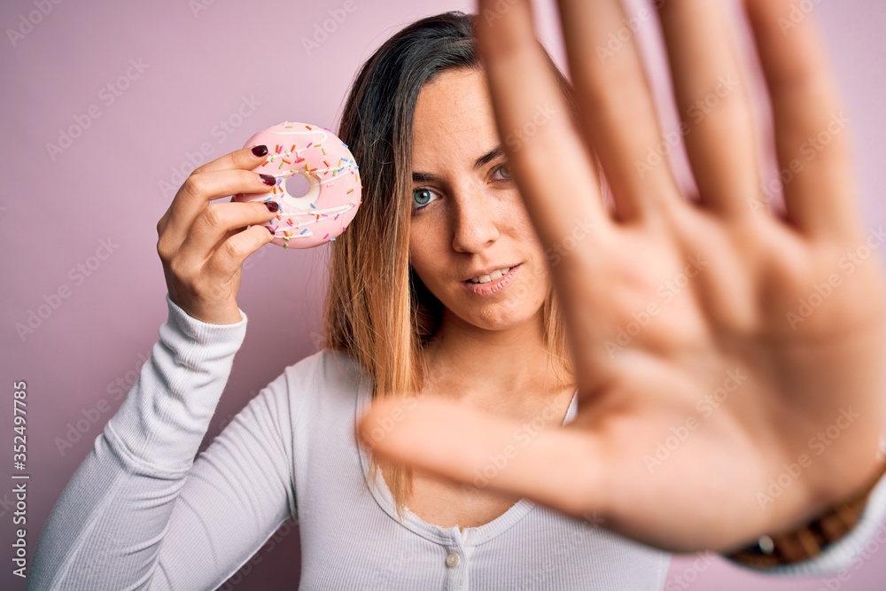 Young beautiful blonde woman with blue eyes holding pink doughnut over isolated background with open hand doing stop sign with serious and confident expression, defense gesture Young beautiful blonde woman with blue eyes holding pink doughnut over isolated background with open hand doing stop sign with serious and confident expression, defense gesture
