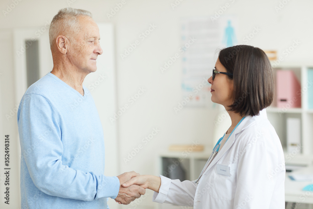 Waist up side view portrait of smiling female doctor shaking hands with senior patient while standing in clinic interior Waist up side view portrait of smiling female doctor shaking hands with senior patient while standing in clinic interior