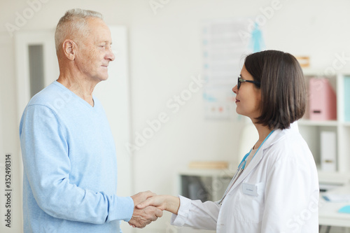 Waist up side view portrait of smiling female doctor shaking hands with senior patient while standing in clinic interior