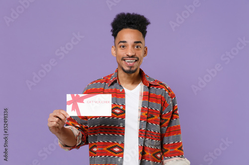 Cheerful young african american guy in casual colorful shirt posing isolated on violet background studio portrait. People sincere emotions lifestyle concept. Mock up copy space. Hold gift certificate.